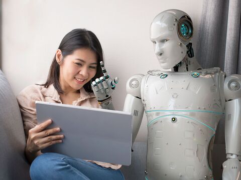 A young woman sitting on a couch, happily interacting with a humanoid robot that is also engaging with a laptop