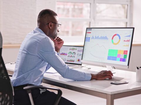 A man in a blue shirt sitting at a desk in an office, analyzing data on a large computer screen displaying various charts, graphs, and data visualizations. Another laptop with similar data visualizations is placed next to the monitor