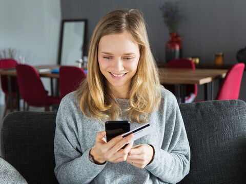 A woman is sitting on a sofa in her room, holding a smart phone and card. She is focused on the phone screen, entering the card's value into an app or website