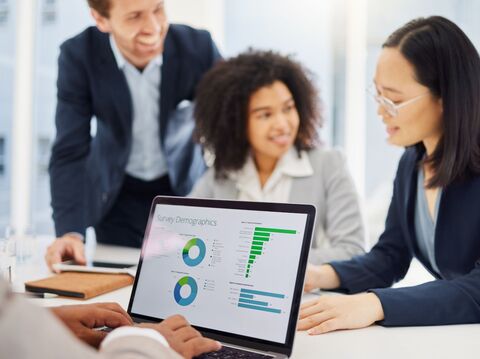 Four business professionals, including two women and two men, are engaged in a meeting around a laptop displaying colorful charts and graphs. One man stands, pointing at the laptop screen, as the team discusses survey demographics. They are in a bright, modern office environment, dressed in formal business attire