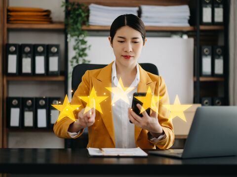 A businesswoman in a yellow blazer using her smartphone in an office setting, with glowing five-star ratings emanating from the device