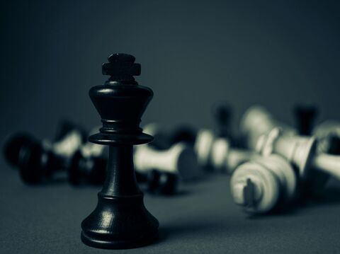 A single black king chess piece standing, with fallen white chess pieces blurred in the background