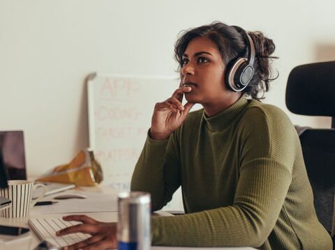 A woman wearing headphones is sitting on a chair near a work desk and thinking about something