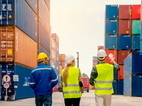 Three logistics workers in safety gear discussing operations at a container shipping yard