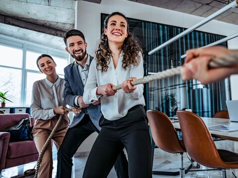 Three office colleagues in a playful tug-of-war, symbolizing teamwork and competition