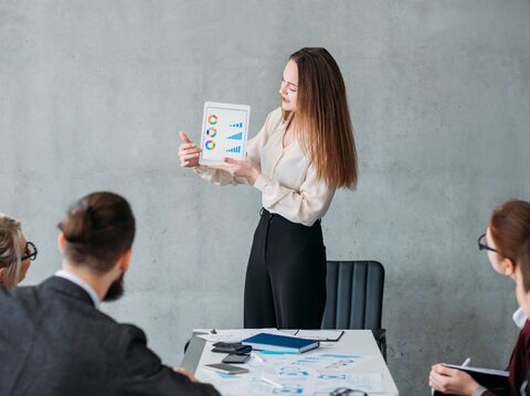 A woman standing in a meeting room and explaining a few graphs in a tablet to some people who are sitting on a chair