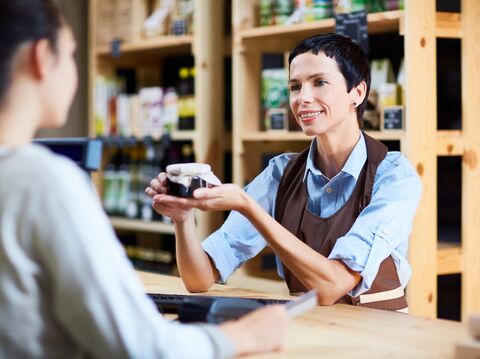 Smiling female clerk in an apron offering a jar of product to a customer at a health food store