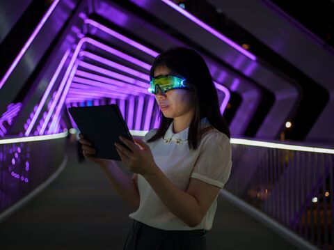 A woman in a modern futuristic setting, wearing light-up smart glasses, intently reads from a tablet. She is on a brightly lit pedestrian bridge at night, illuminated by vibrant purple LED lights that create a dynamic, geometric pattern overhead. Her outfit is business casual, consisting of a white blouse and grey skirt