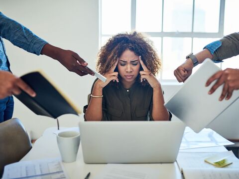 Stressed woman sitting at a laptop, surrounded by colleagues giving her multiple documents