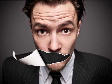 A man in a suit looks directly at the camera with a surprised expression. A piece of paper with a black bar is tearing across his mouth, symbolizing silence or censorship