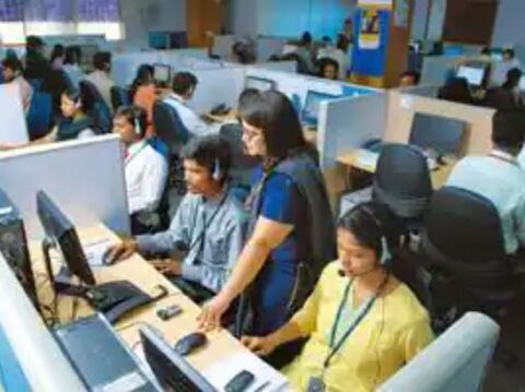 A busy call center with multiple employees working at their computers, some wearing headsets and a supervisor standing and assisting an employee