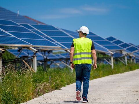 An engineer in a white hard hat and high-visibility vest walking past a large array of solar panels under a clear blue sky