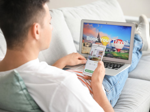 Young man reclining on a sofa, using both a smartphone and a laptop to book travel online, with images of a hot air balloon and landmarks on screens