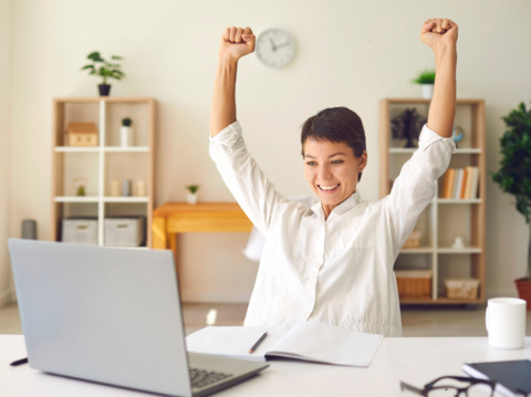 Young woman in a home office raising her arms in victory while sitting at her desk in front of a laptop, smiling joyfully