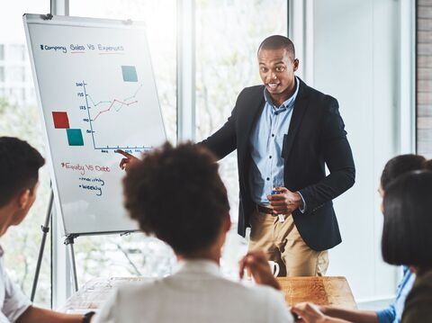 A man in a dark suit is giving a presentation to a group of people. He is pointing at a flipchart displaying a graph titled \"Company Sales VS Expenses\" in a bright office environment with large windows.