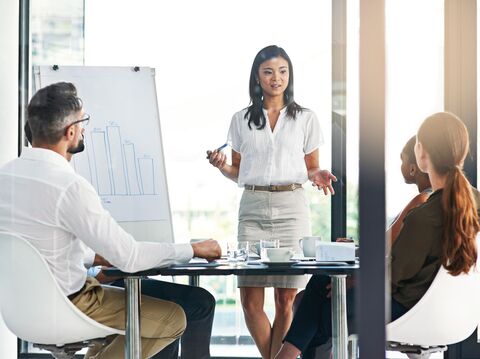 Professional woman giving a business presentation in a conference room, pointing at a flip chart with bar graphs, with attentive colleagues