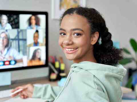 A girl sitting in front of a monitor, smiling and looking backward. Faces on the monitor suggest a video conferencing call