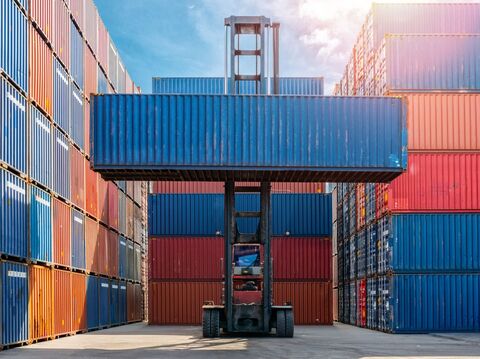 A large, industrial forklift with bright yellow forks raised high in the air, carefully lifting a metal shipping container inside a warehouse