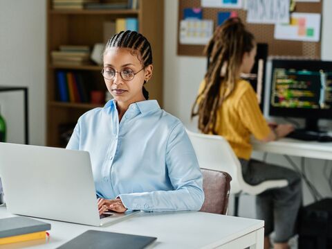 A woman with braided hair, wearing glasses and a light blue shirt, working on a laptop in a modern office environment. In the background, another woman, seated at a desk, is busy with multiple computer screens