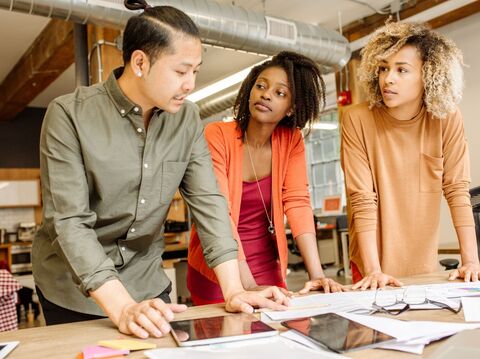 Three colleagues—a man and two women—engaged in a discussion over documents on a table in a creative workspace