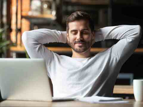 A relaxed young man sits at a table in a cozy café setting, hands clasped behind his head, eyes closed with a content smile on his face. He is wearing a comfortable light gray sweater and is seated in front of a laptop