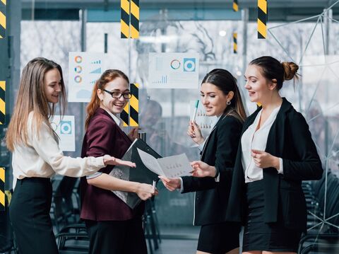 Four women standing in front of a transparent glass covered with few charts and graphs. They are engaged in discussion and analysis of the data presented