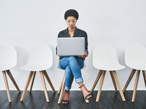 Young woman working on a laptop while sitting alone in a minimalist waiting area with white chairs