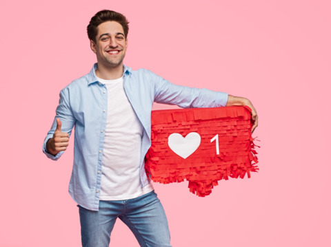 A young man in casual clothing holding a large red 'like' icon, giving a thumbs up, against a pink background