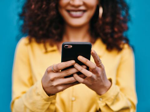 A close-up of a young woman with curly hair, dressed in yellow, happily using her smartphone