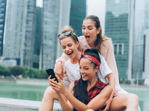 Three women sitting on stairs near a lake, with long buildings in the background. One woman is showing a phone to the others