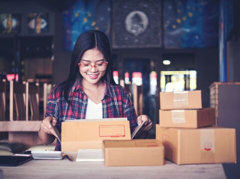 A young woman in a plaid shirt happily opening a cardboard box surrounded by more boxes, indicating a small business setup