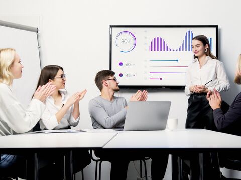 A professional business presentation in a modern office setting. A young woman stands confidently at the front, addressing her colleagues who are seated around a white table. The group, consisting of two men and two women, applauds her presentation displayed on a large screen showing various business metrics and graphs