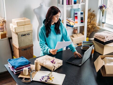 Woman in a teal hoodie reviewing paperwork while packaging orders in a home-based craft studio