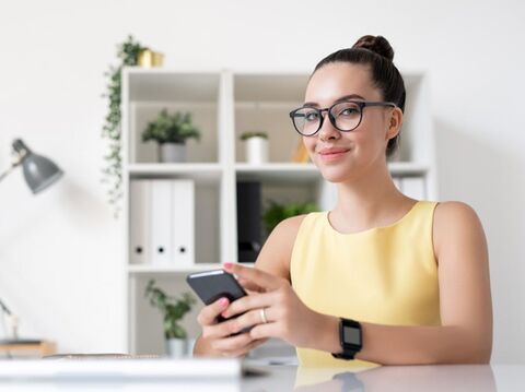 A woman in yellow wearing watch on her right hand and holding phone with both of her hands in front of her torso with a study in her background