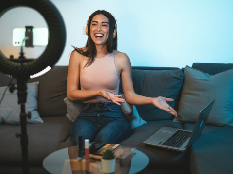 A young smiling woman wearing a light-colored top is sitting on a couch with headphones on, seemingly creating or participating in some audio-related work or activity with recording equipment and a laptop visible nearby