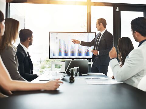 In a modern office setting, a businessperson is giving a presentation using charts or data visualizations displayed on a large screen, while a group of attentive colleagues seated around a conference table observes and takes notes