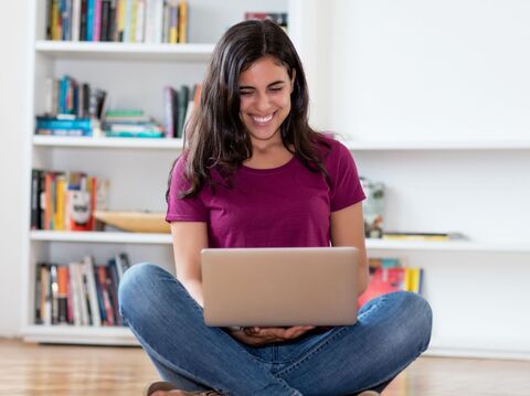 A smiling young woman wearing a purple t-shirt is sitting cross-legged on the floor, working on a laptop surrounded by bookshelves filled with various books and educational materials, in what seems to be a home or study environment