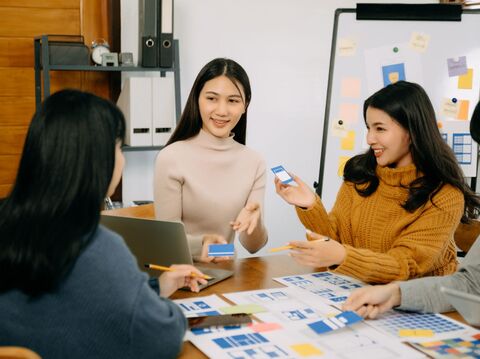 Three people, two young women and one with long dark hair, are collaborating or discussing something while seated around a table or workspace. One woman is holding a smartphone, another is using a laptop, and there are various papers and sticky notes spread out on the table