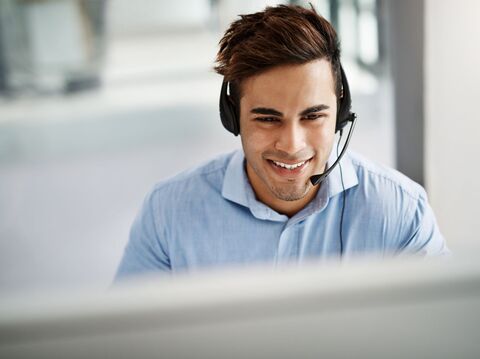 A man in formals wearing a headphone with a microphone sits in front of his laptop with a smiley face