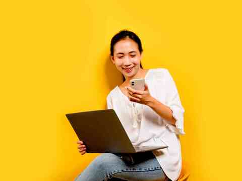 A young Asian woman seated against a vibrant yellow background. She wears a casual white blouse and denim jeans, holding a smartphone in one hand and a laptop in the other