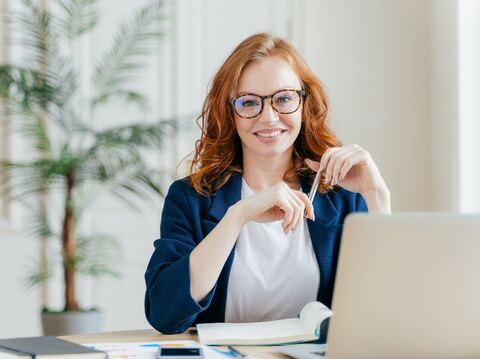 A woman is wearing specs, sitting on a chair in front of a laptop, and holding a pen