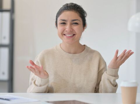 A cheerful young woman in a light beige sweater sits at a white table, engaging in a conversation with an open, welcoming gesture. Her hands are raised slightly with palms facing upwards, and she has a bright, friendly smile