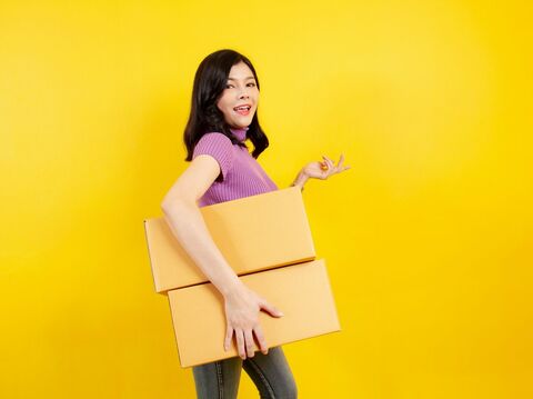 A woman in a purple top holding two cardboard boxes, standing against a bright yellow background