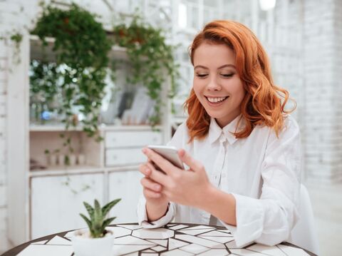 A red-haired woman smiling while looking at her smartphone in a cozy, plant-filled cafe