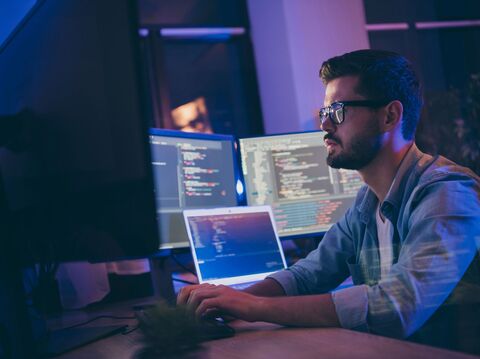 A focused man wearing glasses, working on multiple computer screens displaying code and data