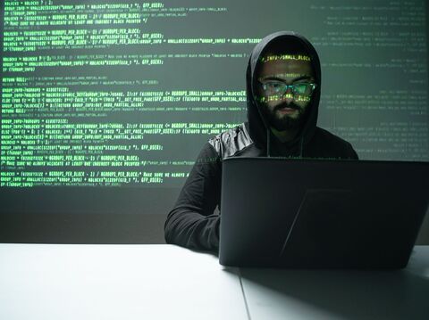 A person in a dark hoodie and glasses working on a laptop with green code projected on a wall behind them