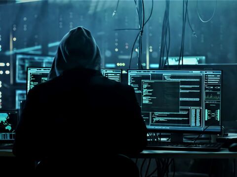 A hooded figure sitting in front of multiple computer monitors in a dark room, depicting a cybersecurity expert or hacker environment with screens showing complex code and data