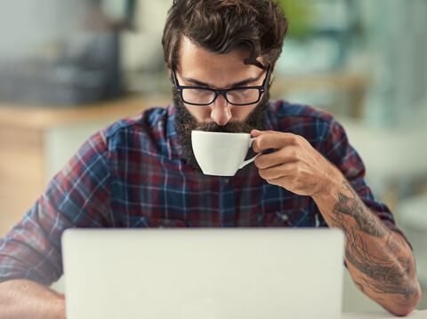 A bearded man wearing glasses and a checkered shirt, sitting at a desk and drinking coffee while working on a laptop. The image represents a tech enthusiast exploring top WordPress plugins