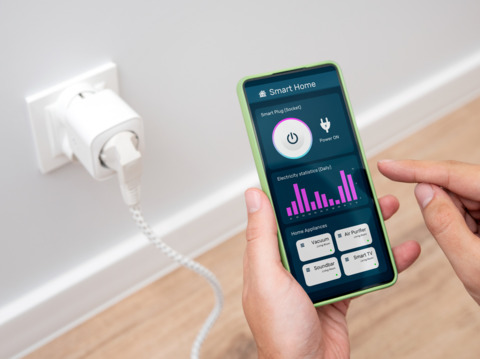 A hand holding a smartphone showing a smart home application, with the phone connected to a smart plug