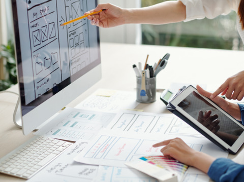 Two people discussing website design, pointing at a screen with wireframe sketches and notes spread out on a desk